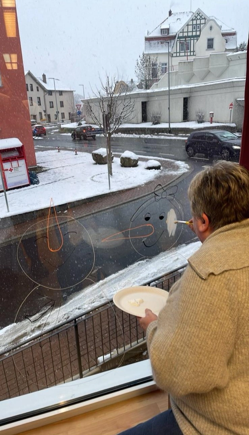 Soziale Teilhabe Kilian und Partner – sozialpädagogische Hilfe – eine Person, die an einem verschneiten Tag Weihnachtsdekorationen wie Schneemänner auf ein Glasfenster malt.