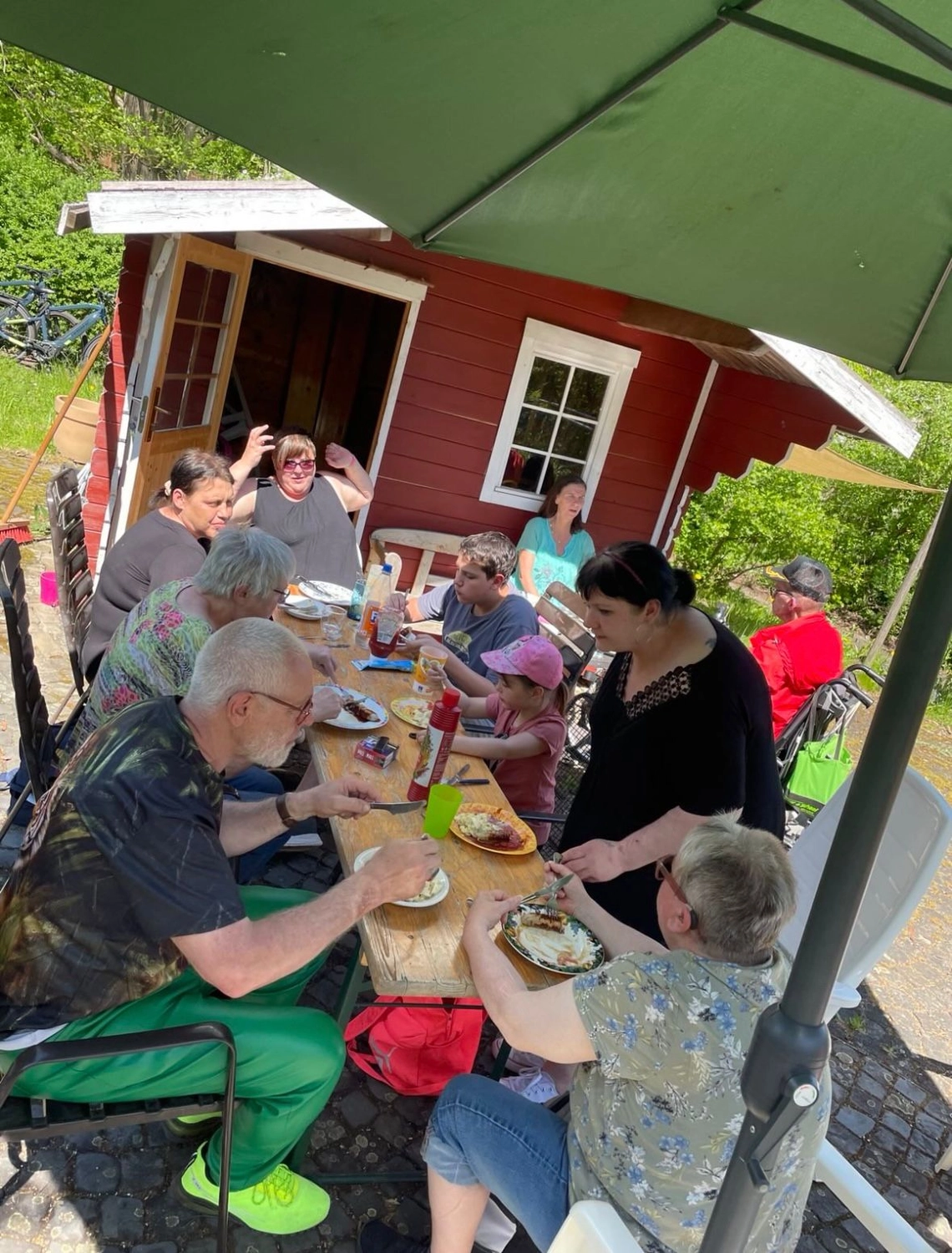 Soziale Teilhabe Kilian und Partner – sozialpädagogische Hilfe – eine Gruppe von Menschen, die vor einer kleinen, traditionellen roten Hütte Essen genießen